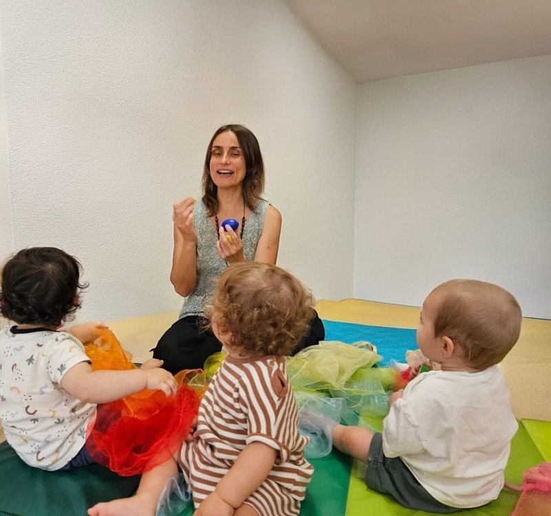 María con tres bebés sentados sobre pañuelos de colores durante una clase de música